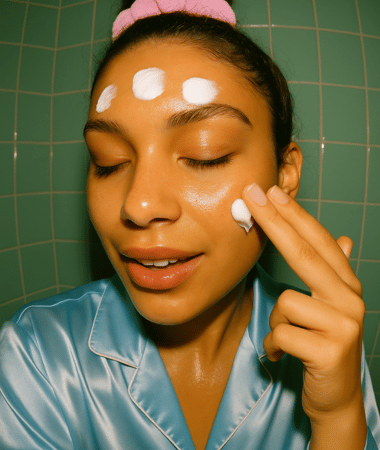 Vier crème-stipjes op voorhoofd; dezelfde vrouw masseert extra product in op wang, satijnen pyjama