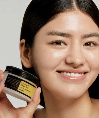 Model met stralende huid glimlacht terwijl ze de COSRX Advanced Snail 92 All in One Cream vasthoudt; benadrukt voedende crème.