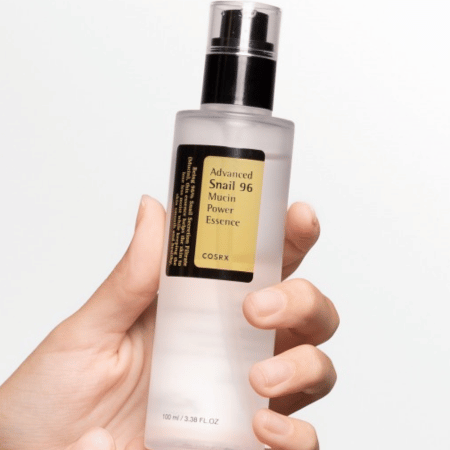 Hand houdt een transparante pompflacon COSRX Advanced Snail 96 Mucin Power Essence omhoog tegen een witte achtergrond.