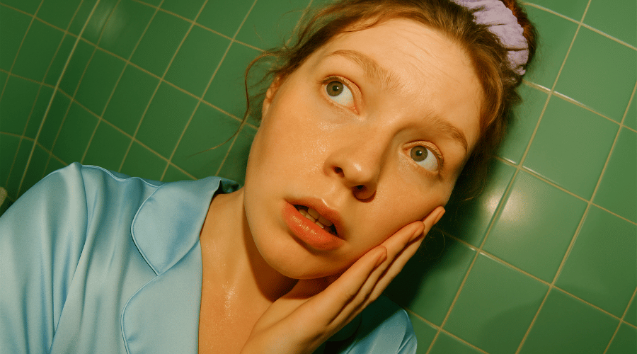 Close‑up van een jonge vrouw die haar gezicht aanraakt in een badkamer met groene tegels; illustreert subtiele fijne lijntjes en rimpels als voorbeeld van huidveroudering.