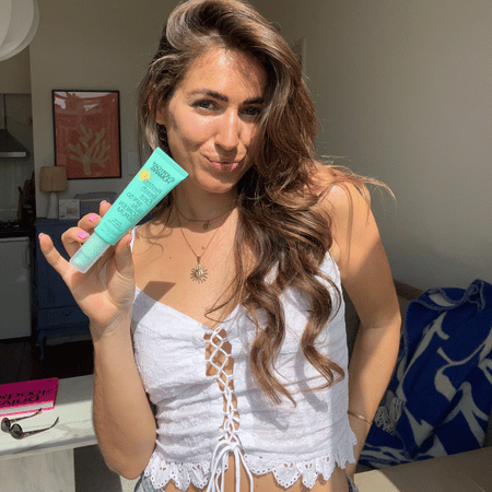 Vrouw met golvend haar in witte top poseert in zonlicht en houdt Everyday Humans Resting Beach Face SPF30 Sun Screen Serum omhoog.