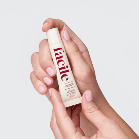 Hand met lichtroze nagellak die een tube Facile Lip Jelly Berry vasthoudt tegen neutrale achtergrond.