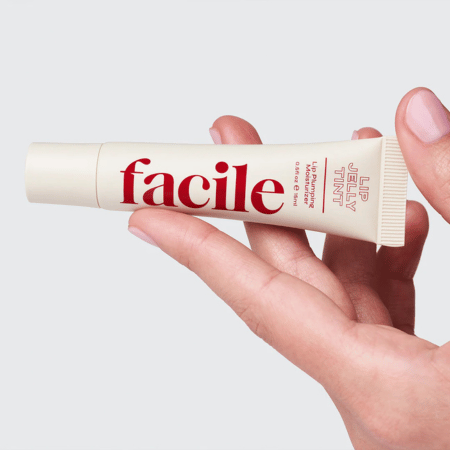 Hand met lichtroze nagellak die een tube Facile Lip Jelly Rouge vasthoudt tegen een zachte, grijze achtergrond.