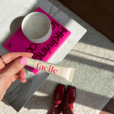 Lifestylefoto waarop iemand een tube Facile Lip Jelly Rose vasthoudt met roze gelakte nagels; op de achtergrond liggen een roze boek en een kaars op een marmeren tafel.