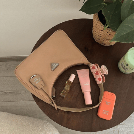 Flatlay van beige Prada‑tas met zomer‑beauty‑essentials: roze zonnebrandcrème, parfumroller, oranje SPF‑case en een bloemen‑haarclip op een houten tafel.