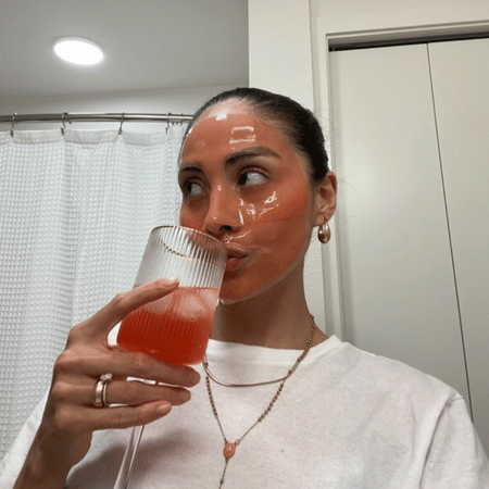Vrouw met glanzend sheet‑masker drinkt een roze mocktail in de badkamer tijdens een self‑care moment.