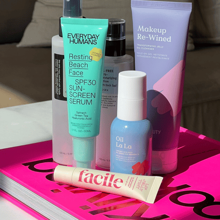Flatlay van kleurrijke huidverzorgingsproducten—waaronder Everyday Humans Resting Beach Face SPF30 serum, Oil La La olie, Facile oogcrème en Makeup Re‑Wined cleanser—op een felroze tijdschrift.