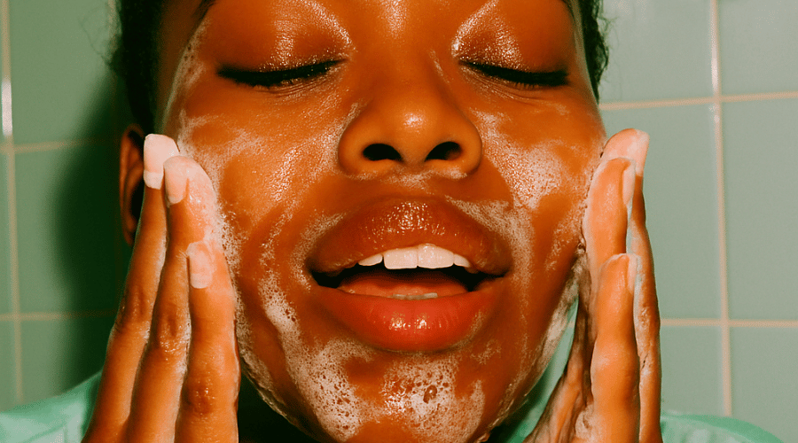 Vrouw masseert een schuimende gel‑cleanser in op haar gezicht tijdens de reinigingsroutine; ogen gesloten, huid glanst zacht.